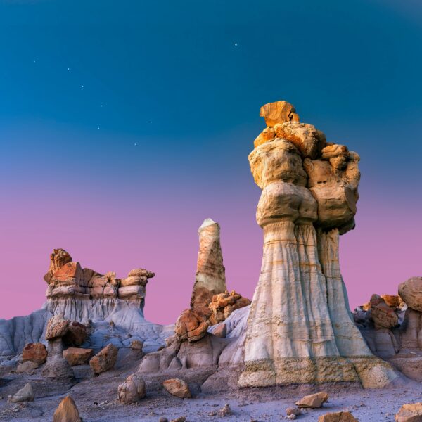 A photo taken in the desert of rock structures, which are called hoodoos, against a gradient blue to purple sky.