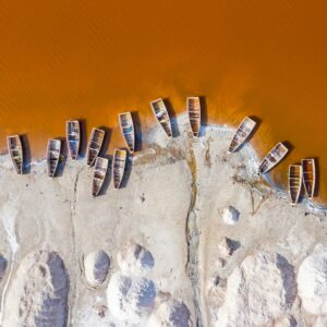 Small boats sit on a shore in West Africa. The water is orange and the beach is a stark white, representing the salt industry.