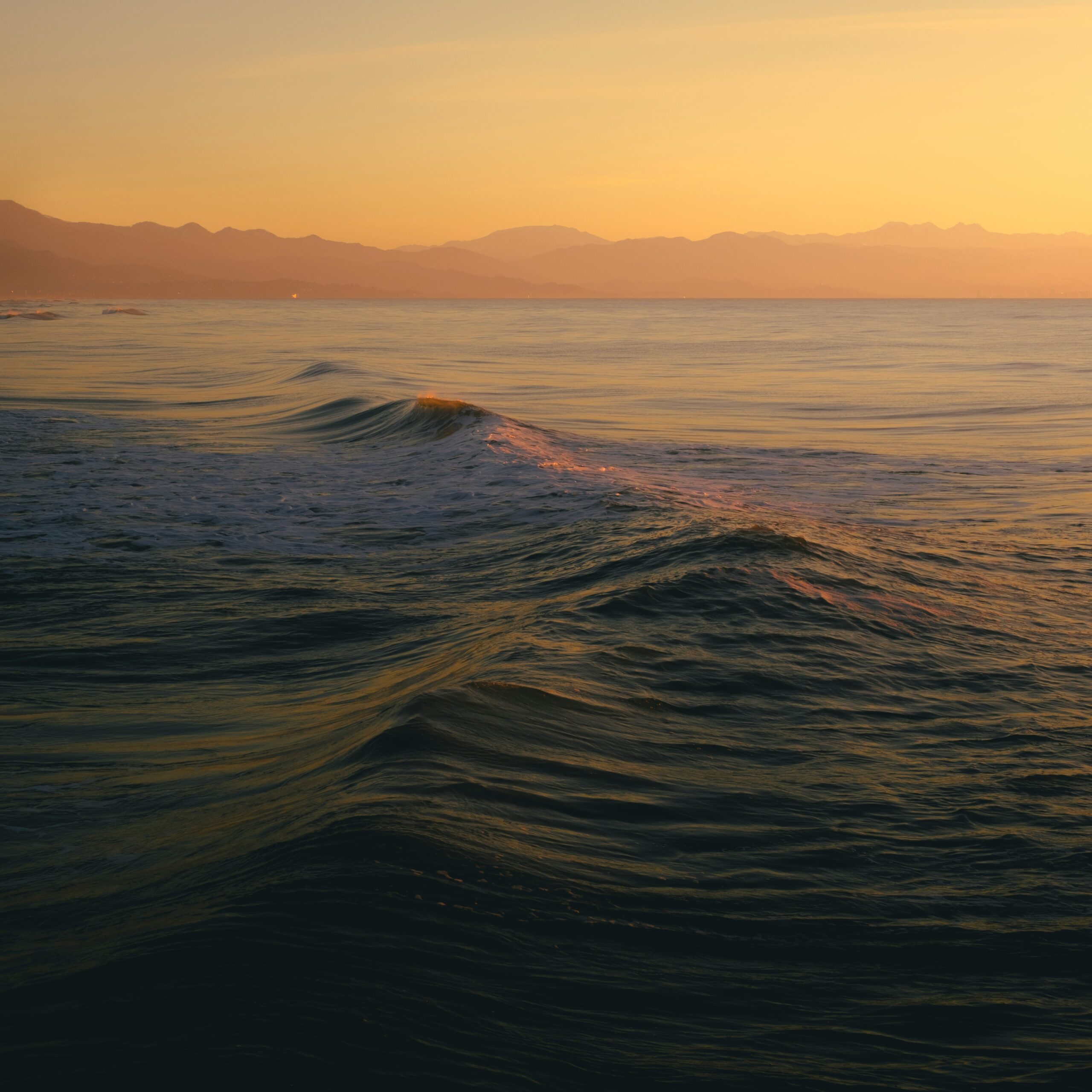 Small waves in the ocean make their way to the shore. The sun is setting in the background, casting an orange glow on the water.