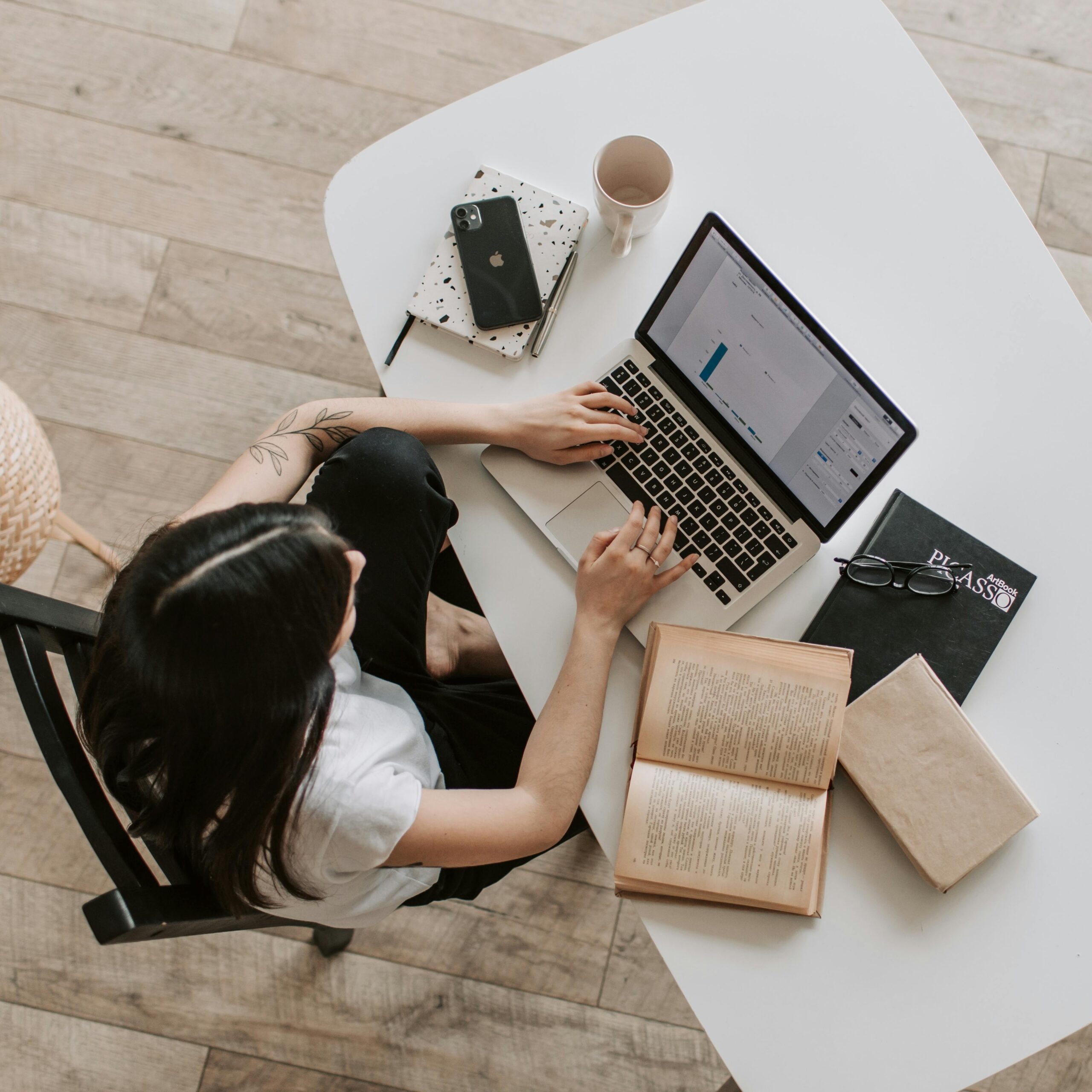 From an aerial perspective, an employee sits at their desk typing on a laptop. Surrounding the laptop are items including books, notebooks, a cell phone, a mug, eye glasses, and a pen.